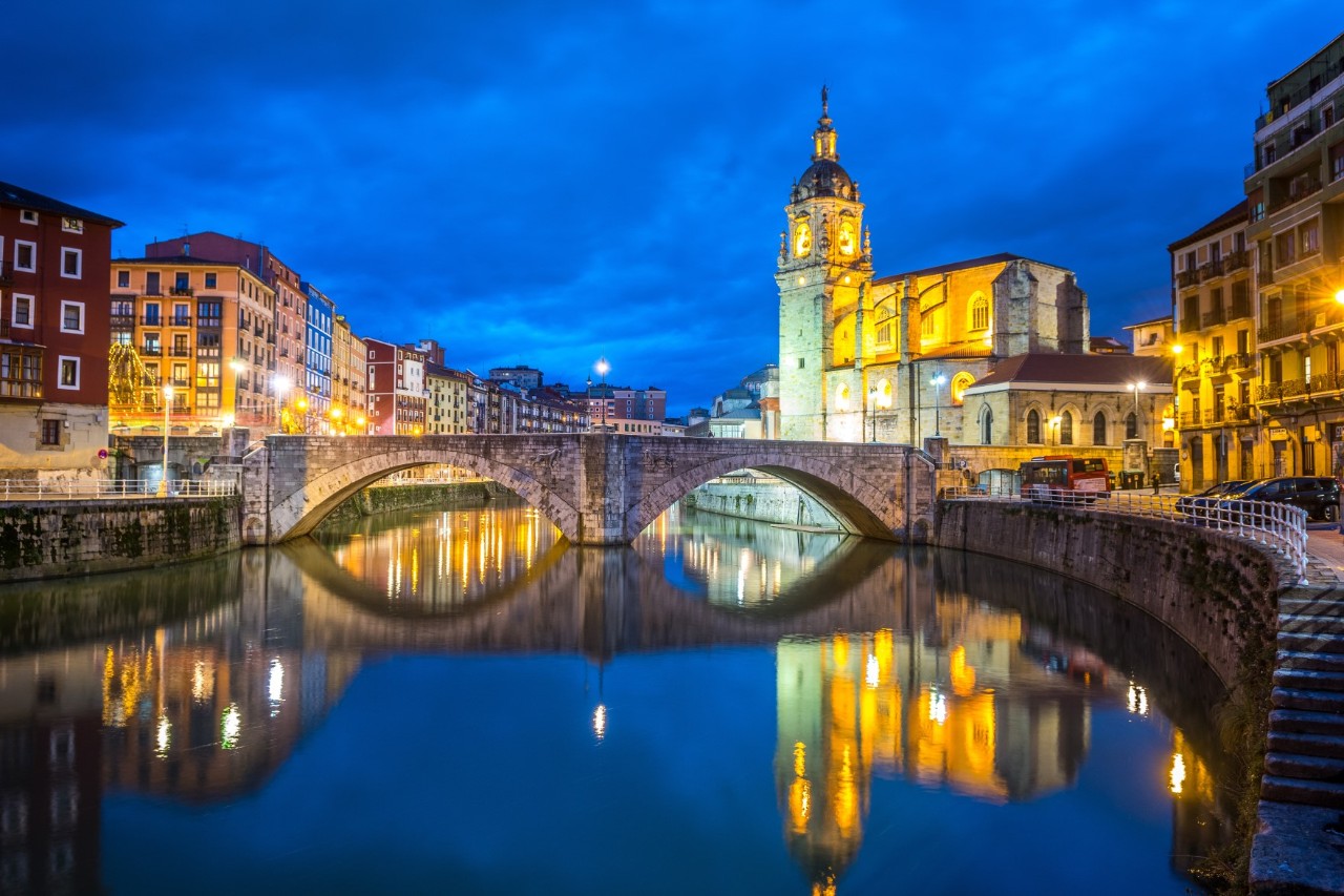 Ene beleuchtete Kirche bei Nacht, die sich im ruhigen Flusswasser spiegelt. Im Vordergrund befindet sich eine alte Steinbrücke, die über den Fluss führt, während sich links bunte Gebäude aneinanderreihen.