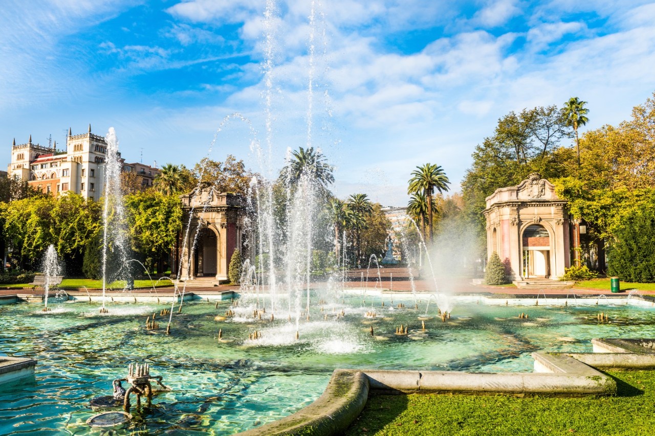 Ein Park mit einem großen Springbrunnen im Vordergrund, umgeben von grünen Bäumen, einer Wiese, Palmen und zwei historischen Pavillons. Im Hintergrund steht ein größeres, weißes Haus. 