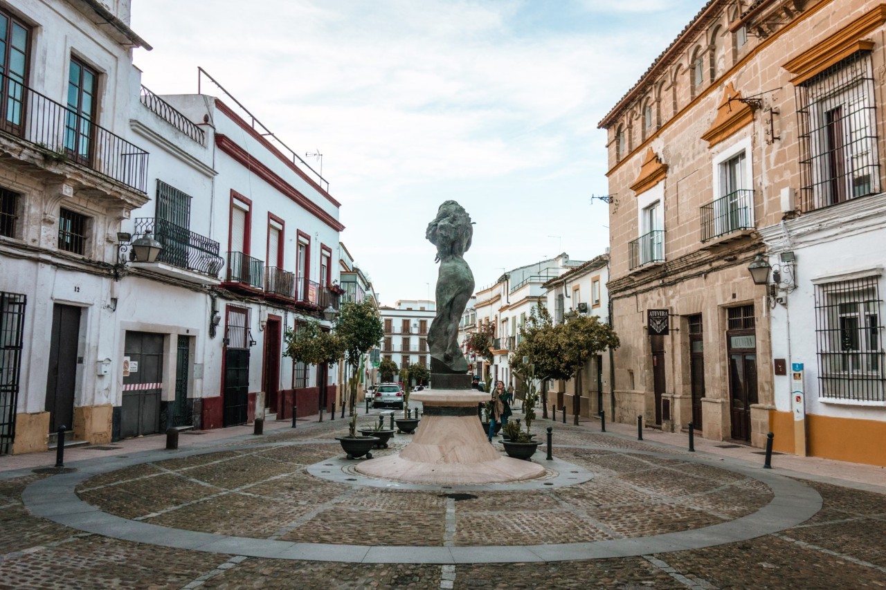 Eine Bronzestatue auf einem runden Platz in einer schmalen Straße. Diese wird von traditionellen andalusischen Gebäuden mit weißen und ockerfarbenen Fassaden umrahmt.