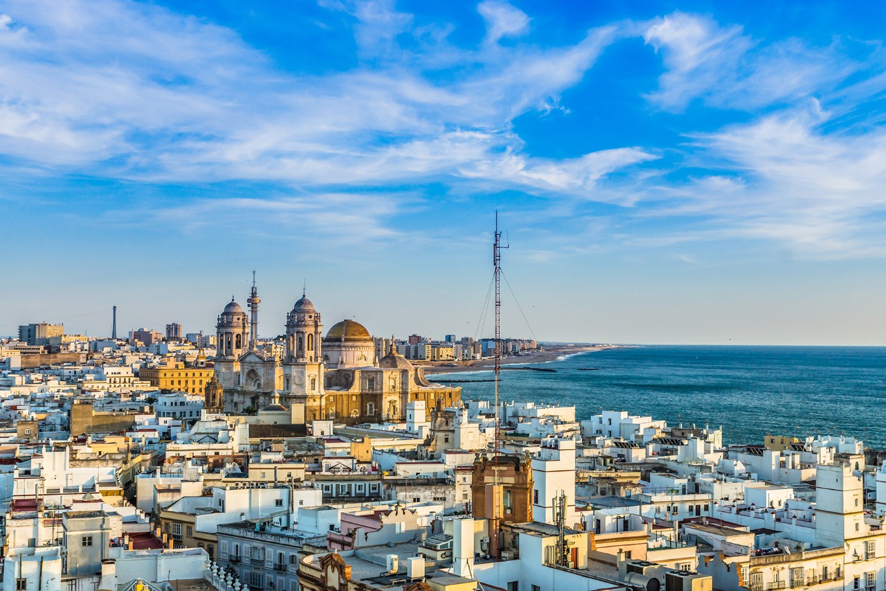 Blick über viele weiße Häuser der Stadt mit einer Kathedrale in der Mitte und dem Meer im Hintergrund unter blauem Himmel.
