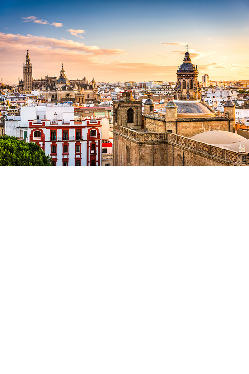 Panoramaaufnahme von Sevilla bei Sonnenuntergang. Im Mittelpunkt stehen eine imposante Kathedrale und der Glockenturm La Giralda. Die Umgebung ist geprägt von weißen Wohnhäusern und historischen Bauwerken. 