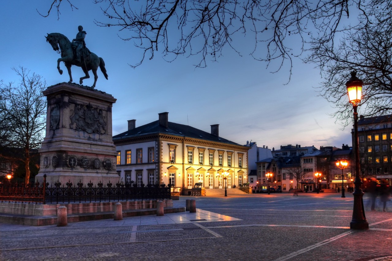 Abendstimmung am menschenleeren Place Guillaume II mit Reiterstatue, Straßenlaternen und beleuchteten Häusern 