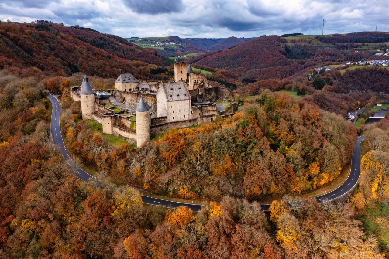 Burg Bourscheid in der hügeligen Landschaft der Ardennen, umgeben vom bunten Herbstwald und einer Straße 