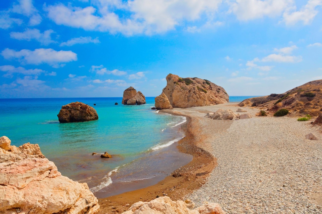 Rechts ist goldener Kieselstrand, auf dem ein großer Felsen platziert ist. Links im Bild sieht man das türkisblaue Wasser, aus dem mehrere Felsblöcke ragen. Der blaue Himmel ist von kleinen Wölkchen durchzogen. 