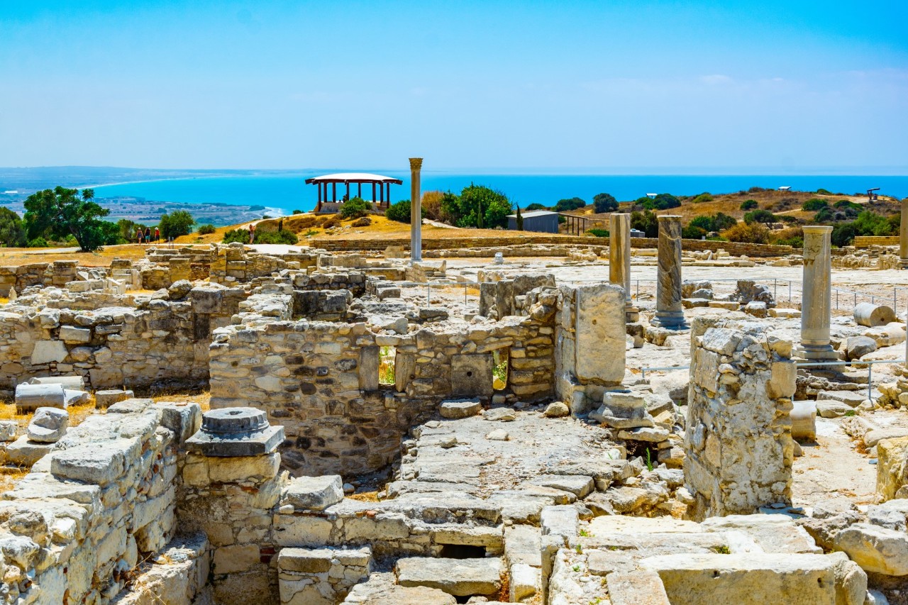 Archäologische Stätte mit zahlreichen Gebäuderuinen, Säulen und ein Tempel mit mehreren Säulen im Bildhintergrund. Dahinter sind mehrere Bäume sichtbar, am Horizont zeichnet sich das Meer ab.