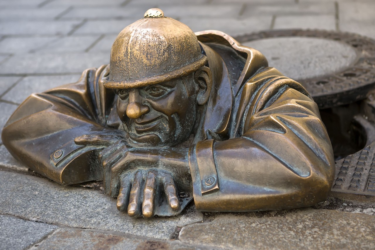 Eine männliche Bronzestatue mit schelmischem Gesichtsausdruck ragt aus einem Kanaldeckel und beobachtet Passanten in der Altstadt.