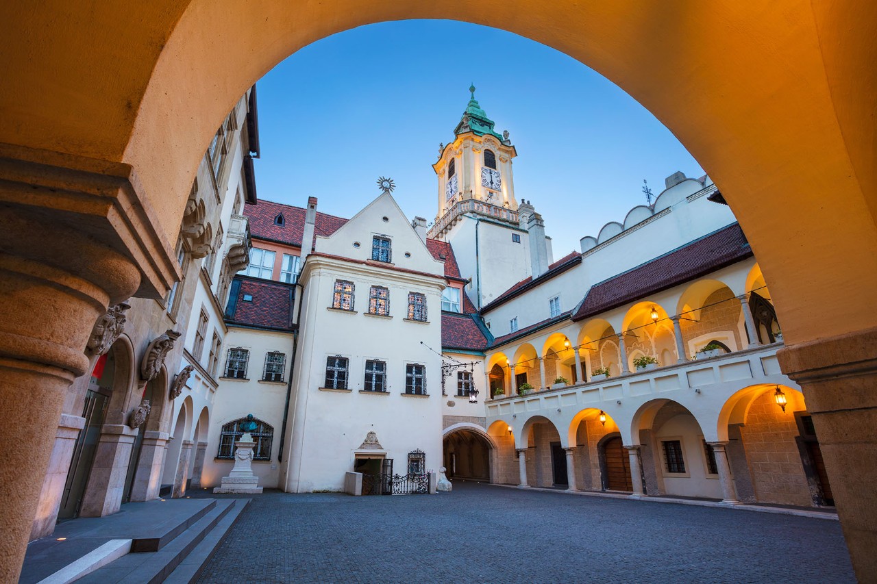 Blick aus einem Torbogen auf ein historisches Gebäude mit markantem Turm, umgeben von Kopfsteinpflaster und alten Häusern.