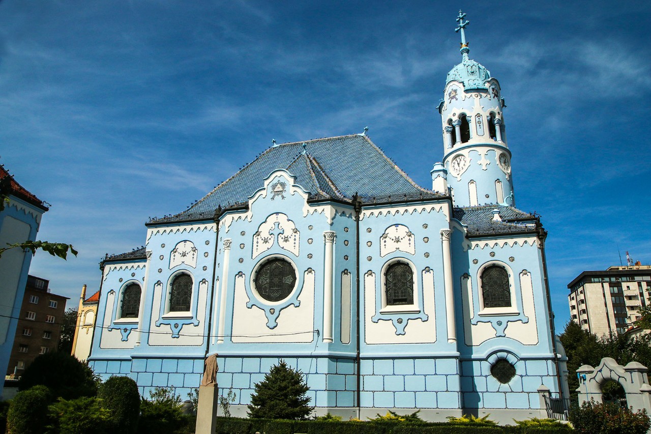 Eine Jugendstil-Kirche mit pastellblauer Fassade, türkis glasiertem Dach und rundem Turm an der rechten Seite.
