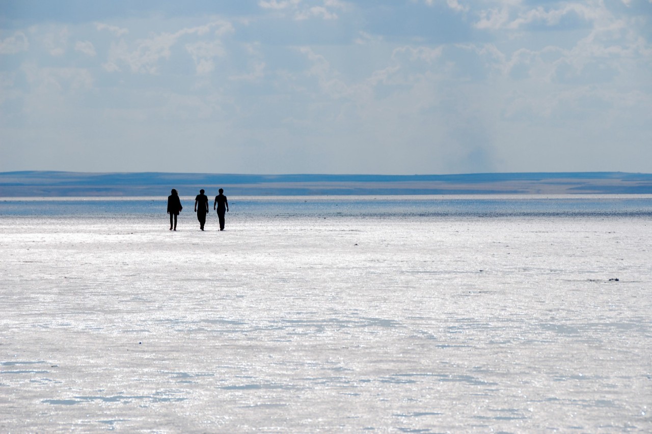 Drei Personen, die als Silhouetten über eine helle, glitzernde Salzfläche gehen. Das dahinterliegende Wasser und der Horizont erstrecken sich weit, der Himmel ist leicht bewölkt.