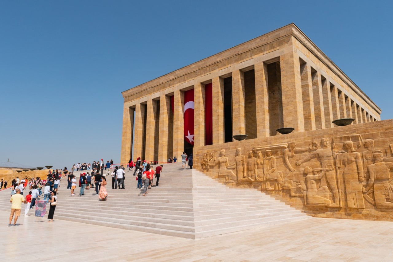 Monumentales Bauwerk mit hohen Säulen aus sand-ockerfarbenem Stein. Auf den breiten Treppen davor sind Besucher zu sehen. An der Fassade des Gebäudes hinter Säulen hängt eine türkische Flagge. Am Treppenaufgang ist ein Relief mit Figuren im antiken Stil eingearbeitet.