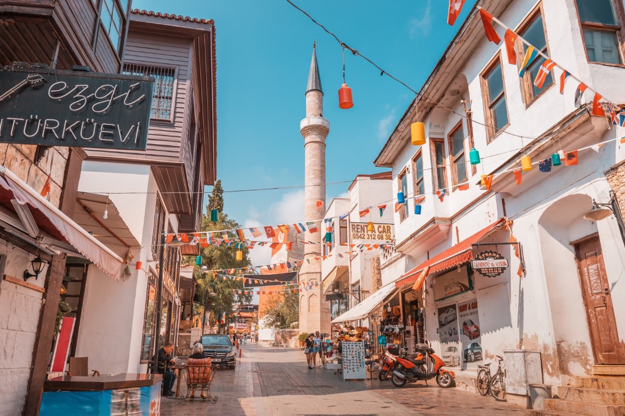 Gasse mit traditionellen Häusern und bunten Wimpelketten, die über die Gasse gespannt sind. Im Hintergrund ist ein Minarett zu sehen. Auf der Straße sind Fußgänger und Geschäfte zu erkennen.