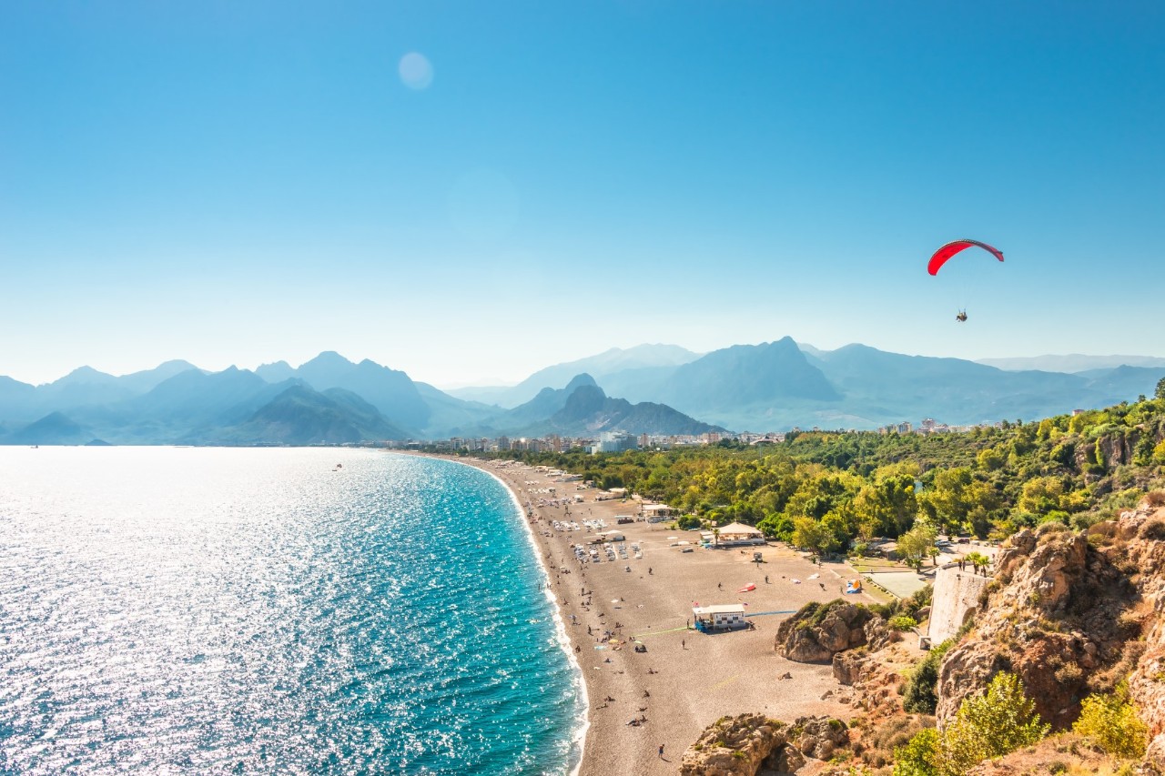 Weitläufiger Strand mit türkisfarbenem Wasser. Im Hintergrund erstrecken sich Berge und grüne Vegetation. Ein Gleitschirmflieger schwebt in der Luft.