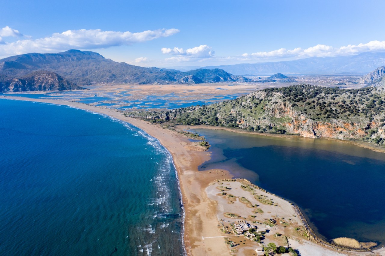 Luftaufnahme eines Naturgebietes. Ein schmaler Sandstreifen trennt das tiefblaue Meer von einer Lagune. Im Hintergrund sind Berge und ein weitläufiges Flussdelta zu sehen. Eine tief liegende Wolkendecke zieht sich über den Himmel.