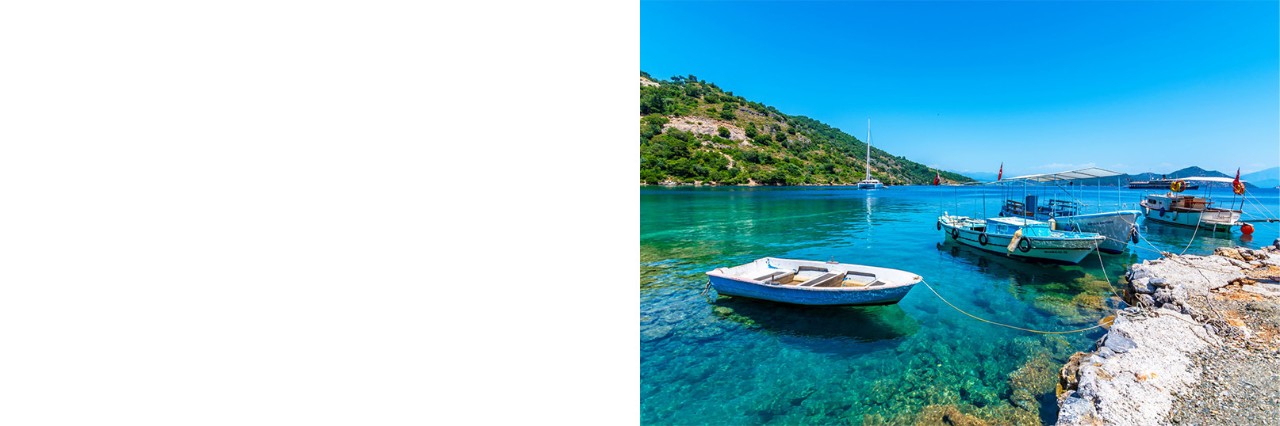 Kleine Boote in kristallklarem, türkisblauem Wasser an einer felsigen Küste, mit bewaldeten Hügeln und einem Segelboot im Hintergrund unter blauem Himmel.