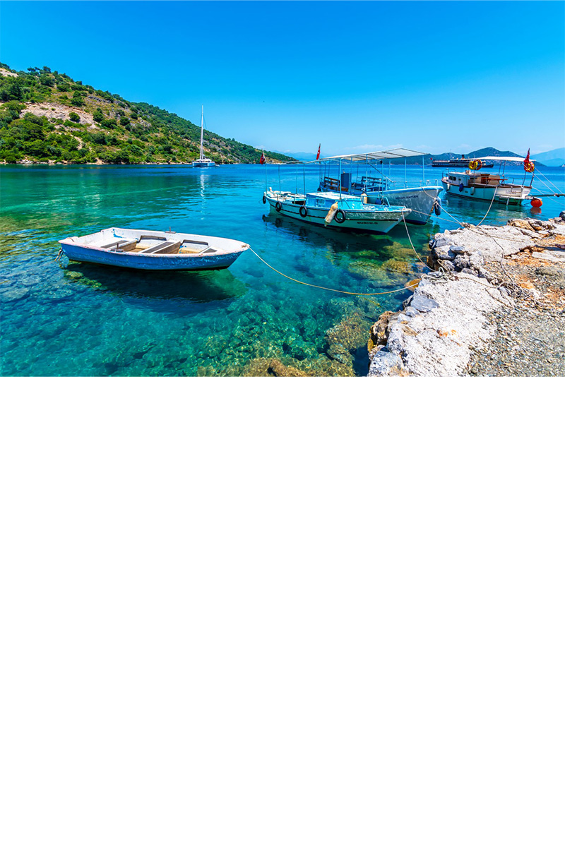 Kleine Boote in kristallklarem, türkisblauem Wasser an einer felsigen Küste, mit bewaldeten Hügeln und einem Segelboot im Hintergrund unter blauem Himmel.