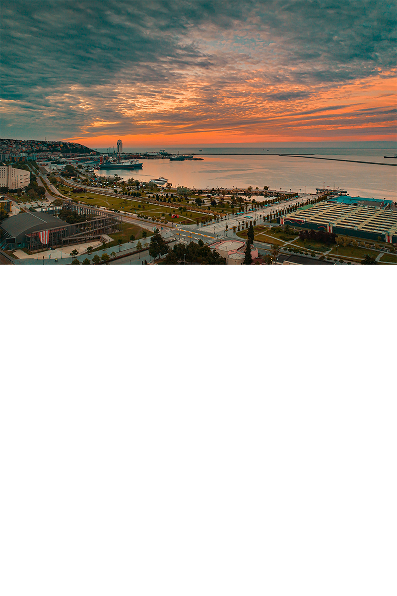 Aufnahme einer Küstenstadt bei Sonnenuntergang mit Park, Häusern, Straßen und einem Hafen mit Schiffen. Der Himmel ist mit orange- und blau gefärbten Wolken bedeckt. 