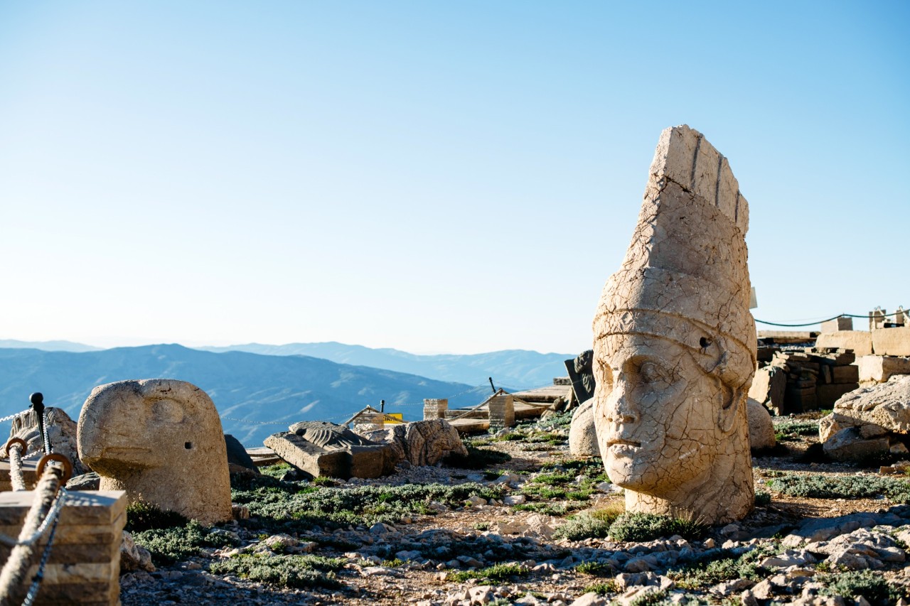 Riesige antike Steinköpfe und vereinzelte Steine mit Blick auf eine weite Berglandschaft.