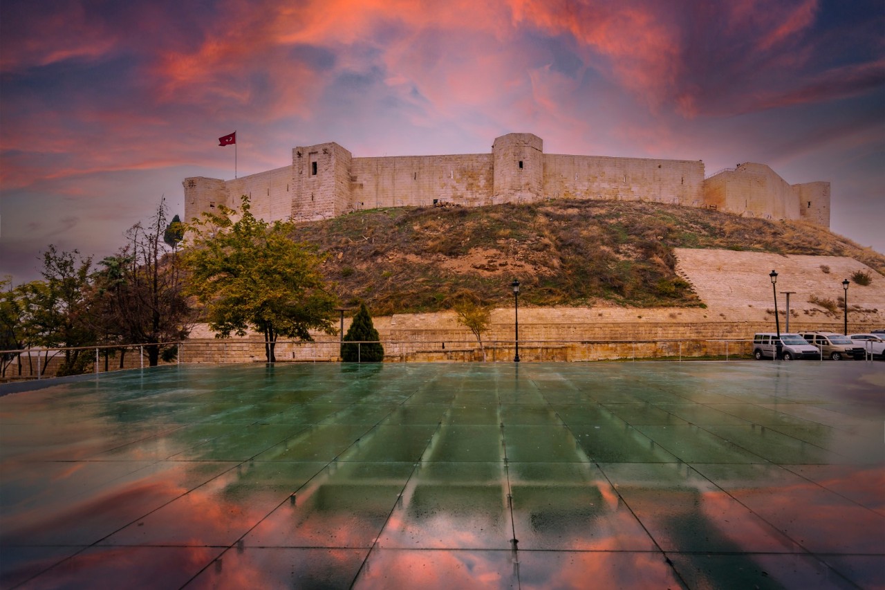 Burg auf einem Hügel bei Sonnenuntergang mit türkischer Flagge, rötlich gefärbtem Himmel und nassem Platz im Vordergrund.