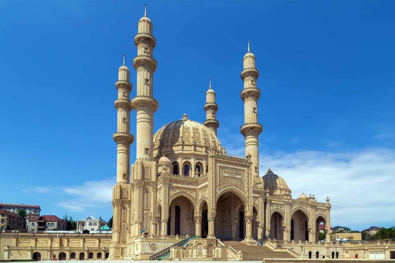 Sandfarbene, riesige Moschee mit großer Kuppel und vier Minaretten. Zahlreiche bogenförmige, verschieden große Tore öffnen sich zur Vorderseite.