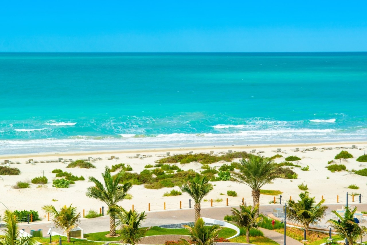 Ein  weißer Sandstrand am türkisblauen Meer. Im Vordergrund sind Palmen und eine schmale Strandpromenade zu sehen. 