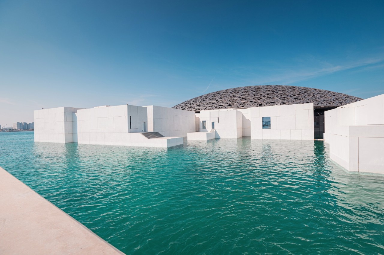 Ein moderner Häuserkomplex mit einer markanten grauen Kuppel und weißen Gebäuden, die von türkisfarbenem Wasser umgeben sind. 