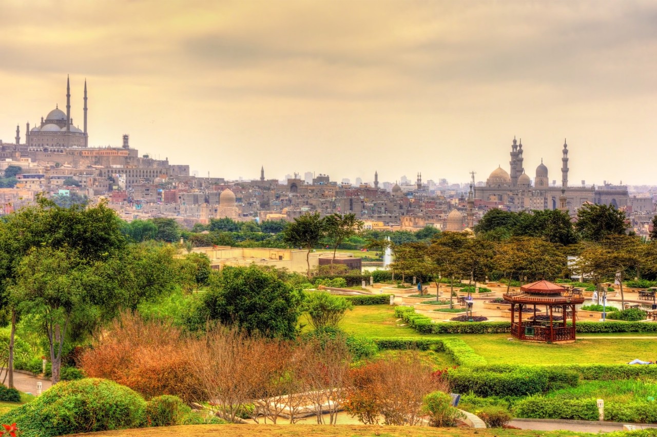 Al-Azhar-Park in Kairo mit gepflegten Grünflächen, Bäumen und einem Pavillon im Vordergrund. Im Hintergrund ist die Skyline Kairos mit mehreren Moscheen und Minaretten unter bewölktem Himmel zu sehen.