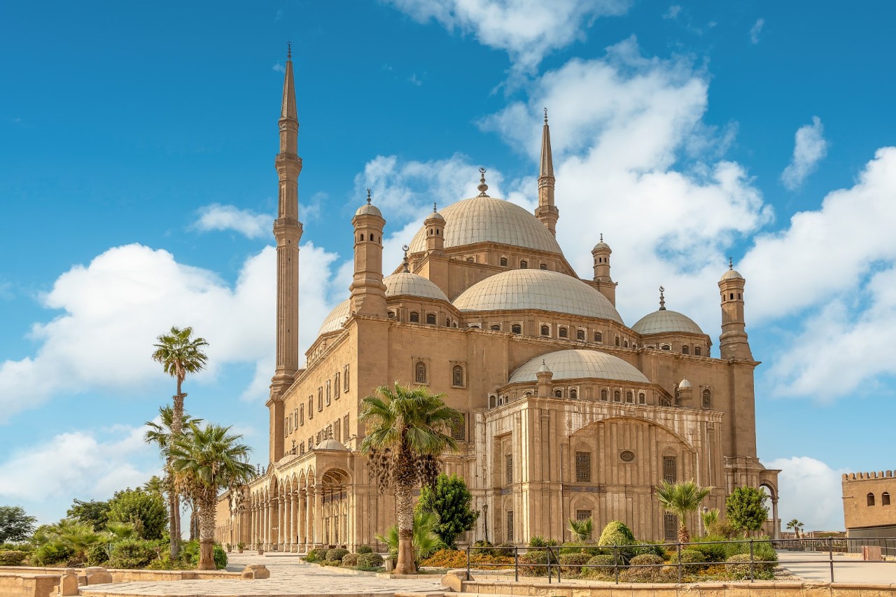 Die Muhammad-Ali-Moschee in Kairo mit zwei Minaretten und mehreren Kuppeln vor blauem Himmel mit wenigen Wolken. Die Moschee ist aus hellem Stein gebaut und von Palmen und Pflanzen umgeben.