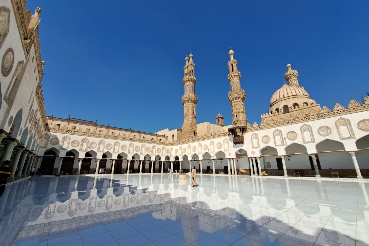 Innenhof der Al-Azhar-Moschee in Kairo mit glänzendem, hellen Boden und umlaufenden Arkaden. Zwei Minarette und eine große Kuppel ragen vor klarem, blauem Himmel über das Gebäude hinaus.