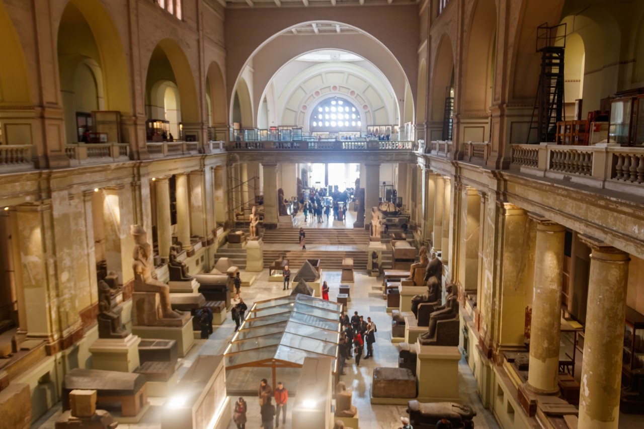 Innenansicht des Ägyptischen Museums in Kairo mit hohen Säulen, Ausstellungsstücken wie Statuen und Sarkophagen sowie Besuchergruppen im Raum. Tageslicht fällt durch ein großes Fenster am Ende der Halle.
