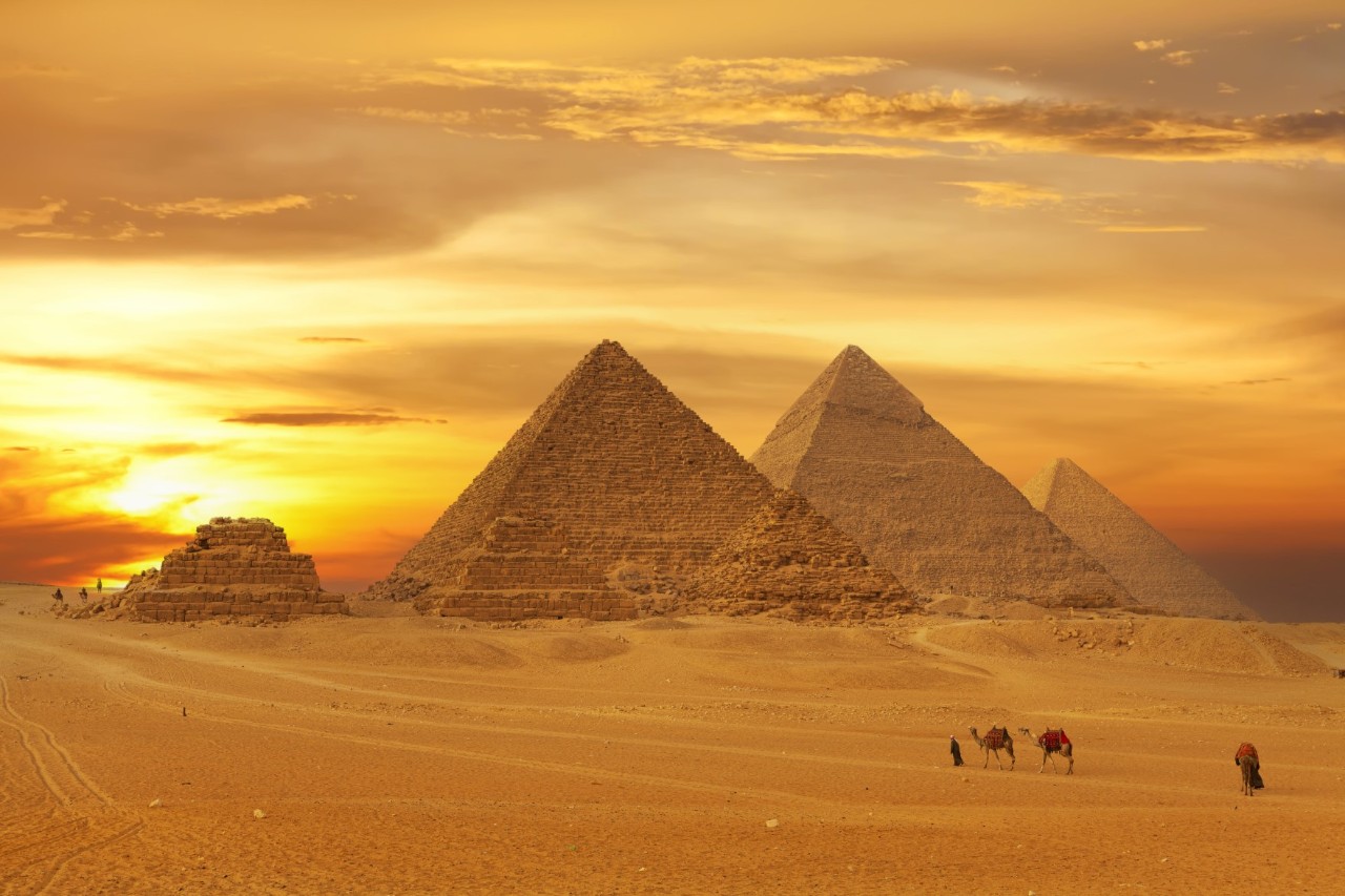 Die Pyramiden von Gizeh bei Sonnenuntergang mit orangem Himmel im Hintergrund. Im Vordergrund befinden sich Wüste und mehrere Kamele mit Reitern.