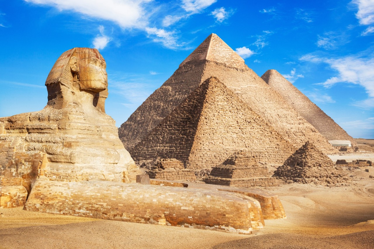 Die Große Sphinx von Gizeh in Seitenansicht mit den drei Pyramiden im Hintergrund unter klarem, blauem Himmel. Die Szene zeigt eine Wüstenlandschaft mit antiken Bauwerken.