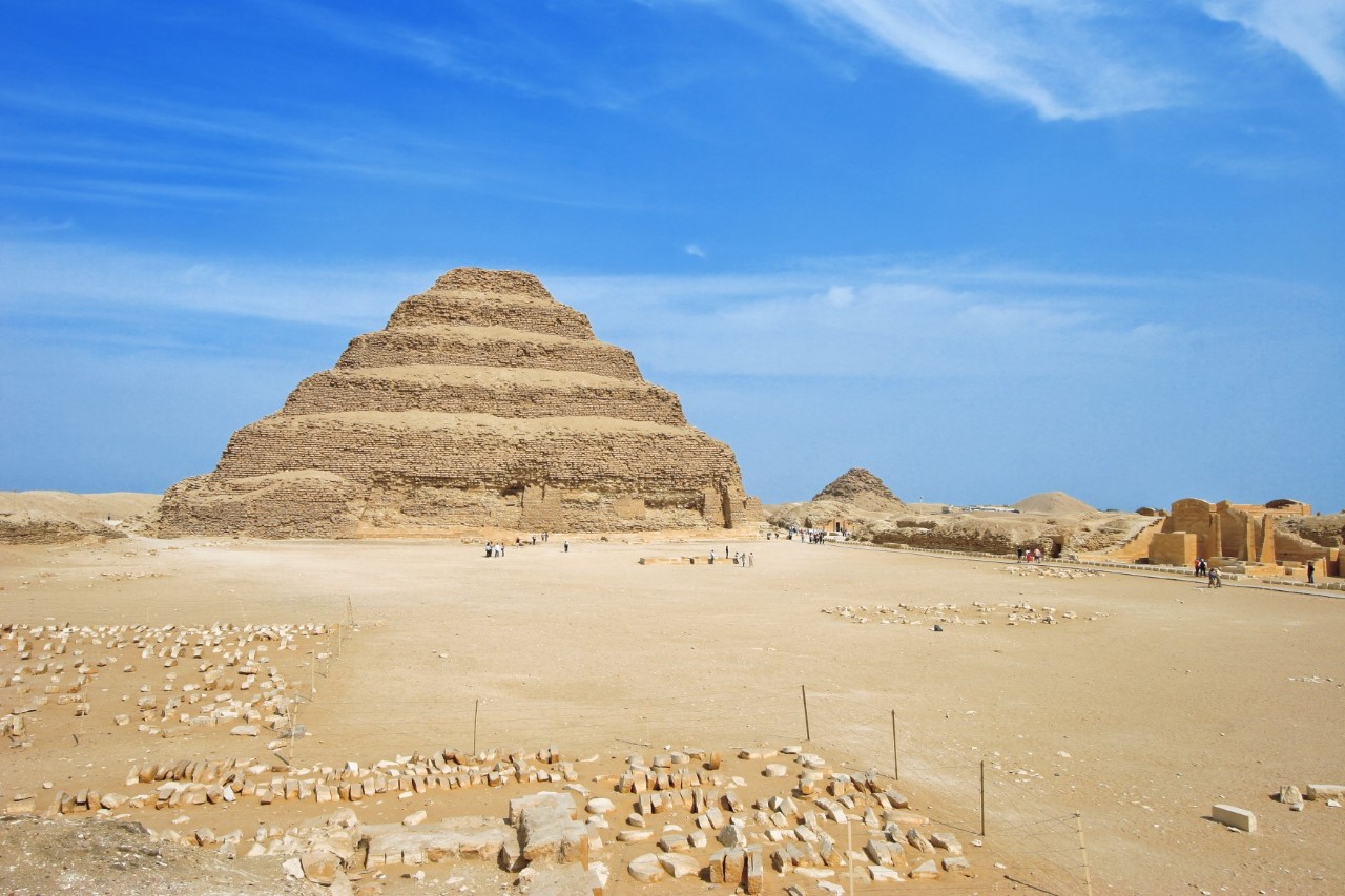 Die Stufenpyramide von Sakkara in Ägypten vor blauem Himmel. Die Pyramide steht in einer weitläufigen Wüstenlandschaft mit verstreuten Steinen und kleineren Ruinen im Vorder- und Hintergrund.