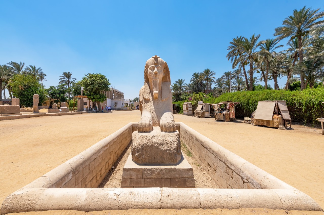 Steinerne Alabaster-Sphinx von Memphis in Ägypten, frontal aufgenommen, stehend in einer rechteckigen Umfassung. Im Hintergrund sind Palmen, Bäume und kleine Gebäude unter blauem Himmel zu sehen.