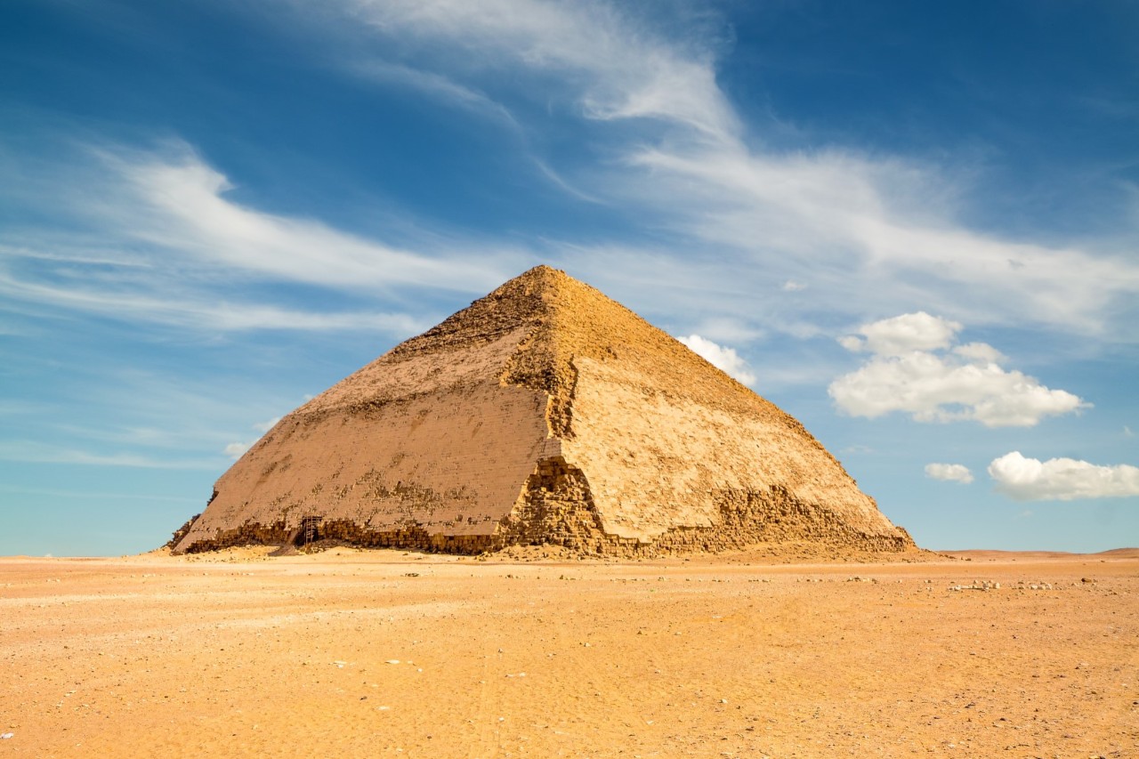 Die Knickpyramide von Dahschur in Ägypten steht in einer offenen Wüstenlandschaft unter blauem Himmel mit vereinzelten Wolken. Die Pyramide zeigt einen Knick in der Neigung ihrer Seitenflächen.