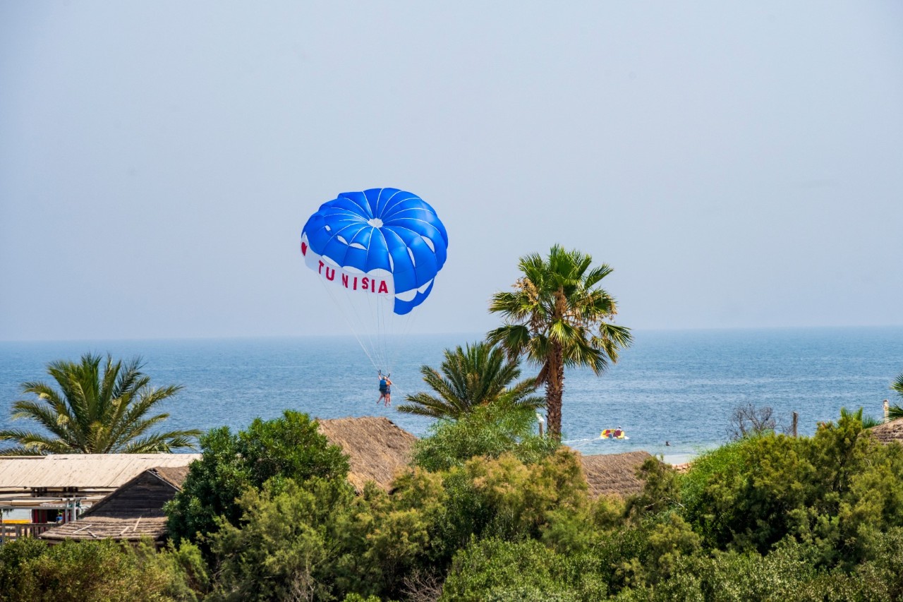 Person beim Parasailing, mit blauem Fallschirm, auf dem das Wort „Tunisia“ steht, über dem Meer. Im Vordergrund sind Palmen und grüne Vegetation zu sehen. 