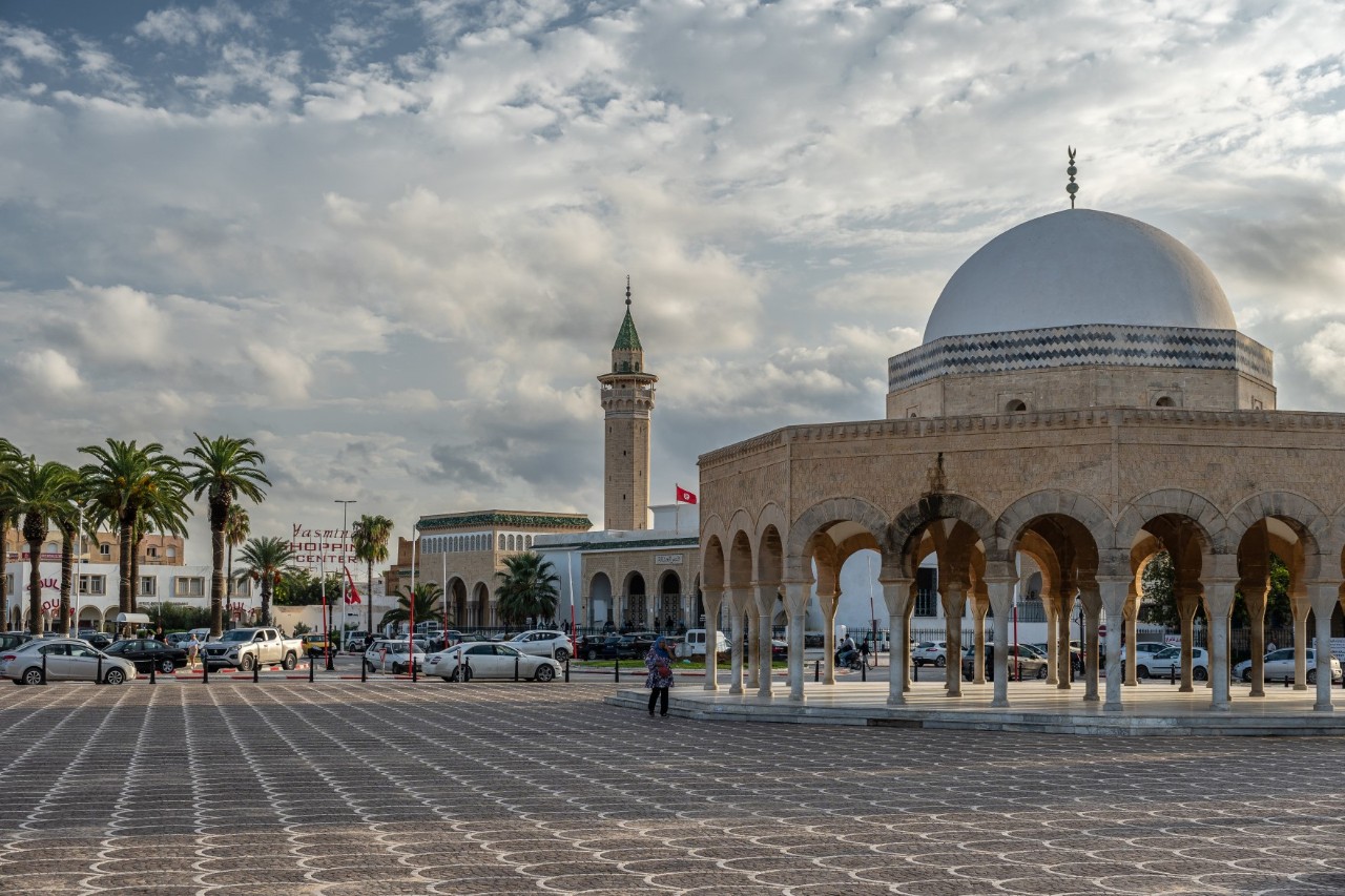 Auf einem städtischen gepflasterten Platz. Im Vordergrund steht eine große Kuppel mit darunter liegenden verzierten Bögen. Daneben befindet sich ein Minarett. Im Hintergrund sind Palmen, Autos und Häuser zu sehen. 