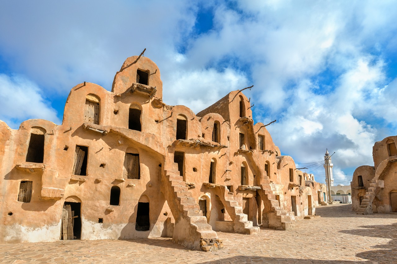 Mehrstöckige, sandfarbene Lehmgebäude mit hölzernen, teils halbrunden Türen und Außentreppen in einem traditionellen Wüstendorf unter blauem Himmel mit Wolken.