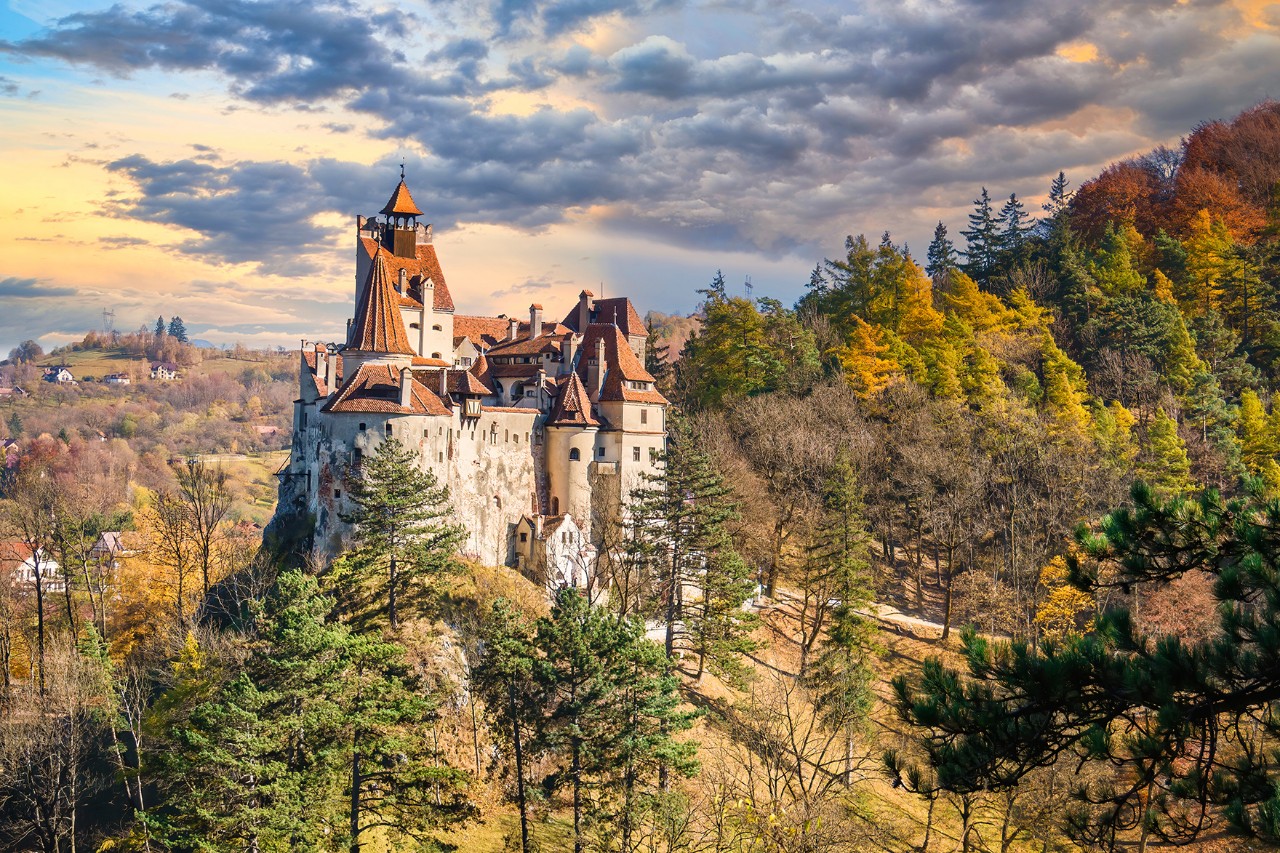 Das Bild zeigt ein Schloss mit unterschiedlichen Türmen und roten Dächern auf einem Hügel. Drumherum befinden sich viele herbstlich bunt eingefärbte Bäume.