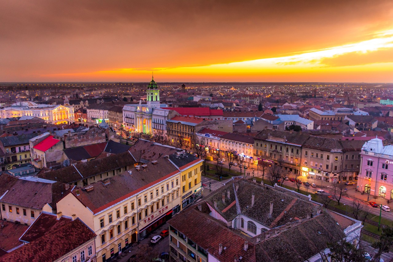Luftaufnahme einer Altstadt bei Sonnenuntergang mit beleuchtetem Kirchturm und orangefarbenem Himmel.