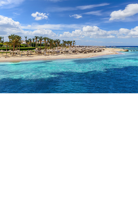 Sandstrand mit klarem, türkisfarbenem Wasser, Palmen im Hintergrund und mehreren Sonnenschirmen. Ein Steg führt ins Meer. 