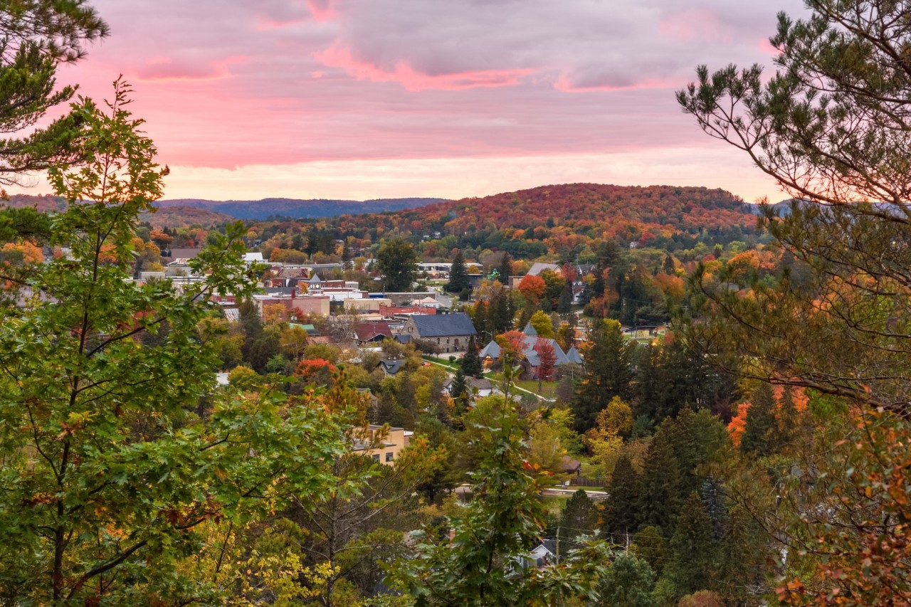 Herbstliche Kleinstadt inmitten einer hügeligen Landschaft. Bunte Laubbäume prägen das Panorama, während der Himmel in Rosa- und Orangetönen erstrahlt.