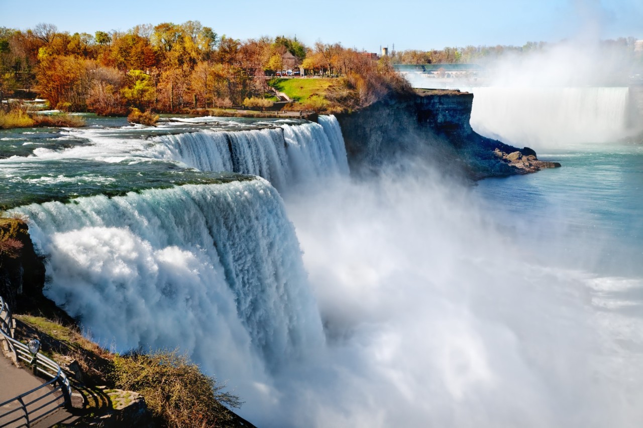 Wasserfälle, bei denen mächtige Wassermassen tosend in die Tiefe stürzen, umgeben von dichter Gischt und einer herbstlich gefärbten Baumlandschaft.