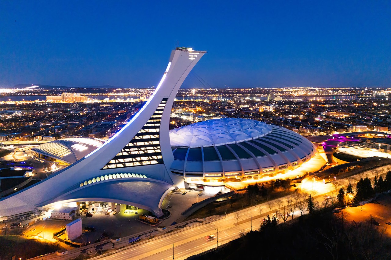 Nächtliche Aufnahme eines Stadions mit einem schrägen, futuristisch aussehenden Turm. Im Vordergrund ist eine beleuchtete Straße und im Hintergrund sind die Lichter der Stadt vor dunkelblauem Himmel zusehen.