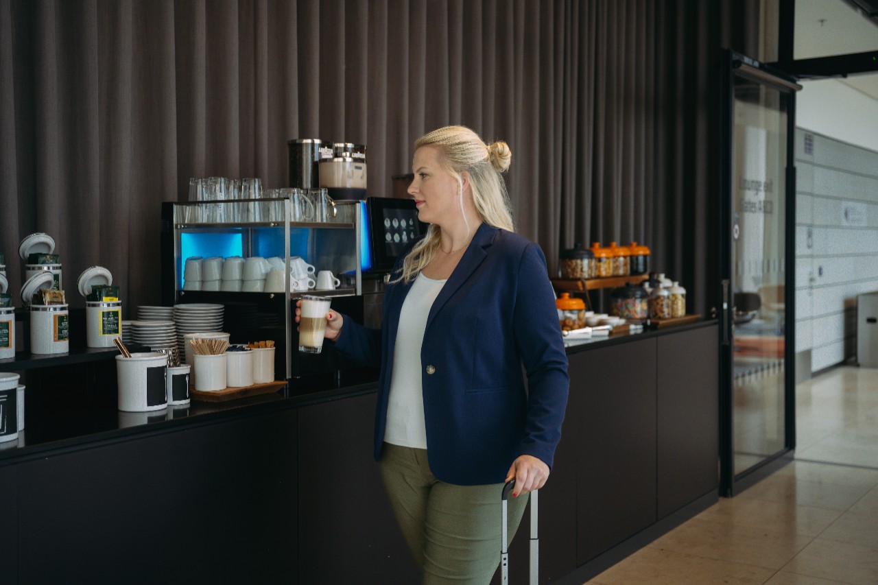 Eine blonde Frau geht mit Koffer in der einen Hand und Kaffee in der anderen Hand an einem Buffet entlang.