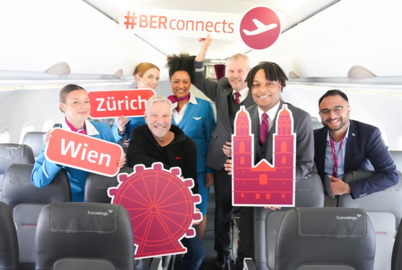 Eine Eurowings-Crew und weitere Personen in einem Flugzeug, alle lächeln in die Kamera und halten Schilder in den Händen. Darauf steht "#BER connects", "Zürich", "Wien". Zwei weitere Schilder zeigen Wahrzeichen von Wien und Zürich. 
