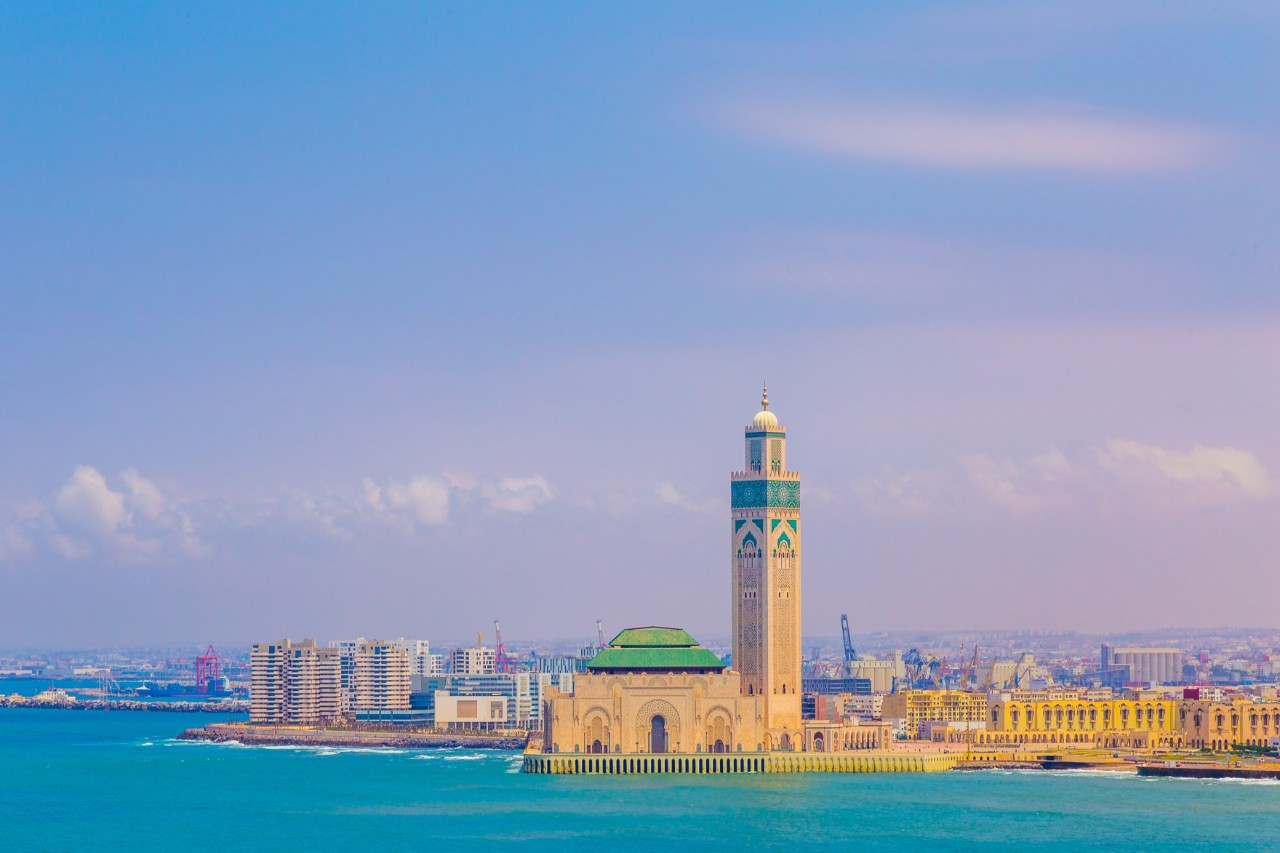 Weitläufiger Blick über das blaue Meer auf eine Stadt mit zahlreichen Häusern. Zentral im Bild steht eine große Moschee mit einem hohen, eckigen Turm. Der Himmel ist lilafarben und leicht diesig.