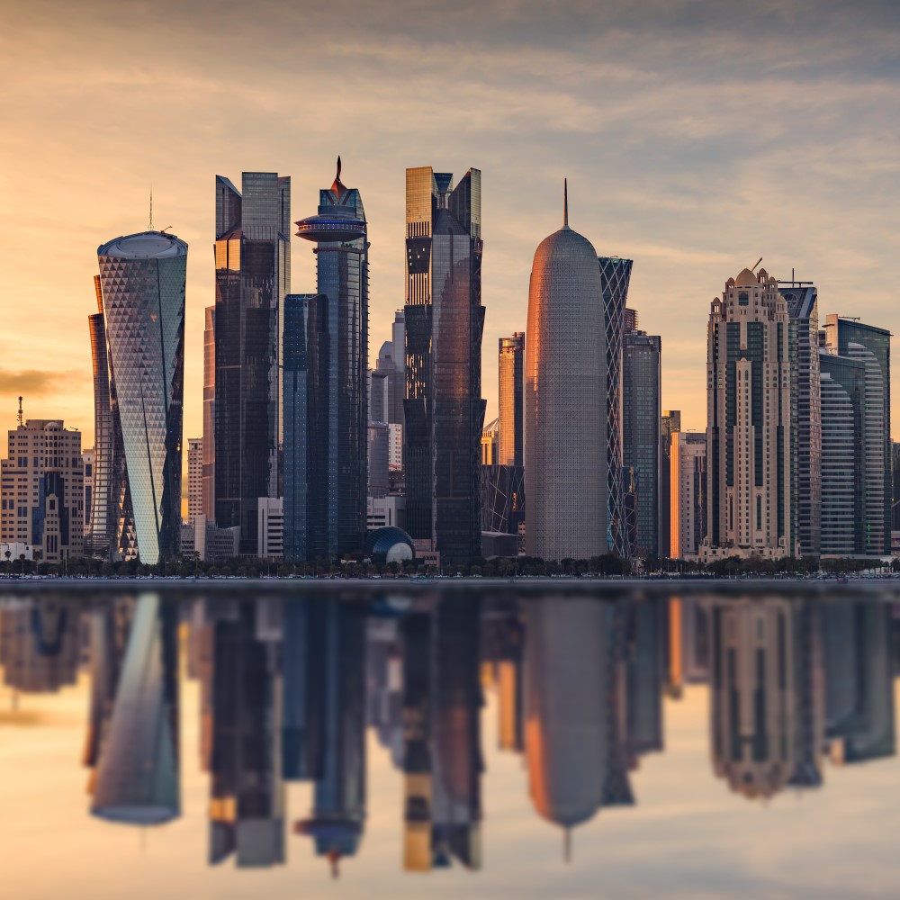 Skyline der Stadt Doha mit einem Gewässer im Vordergrund. Die Szenerie wird in ein orangenes Licht von der untergehenden Sonne getaucht.