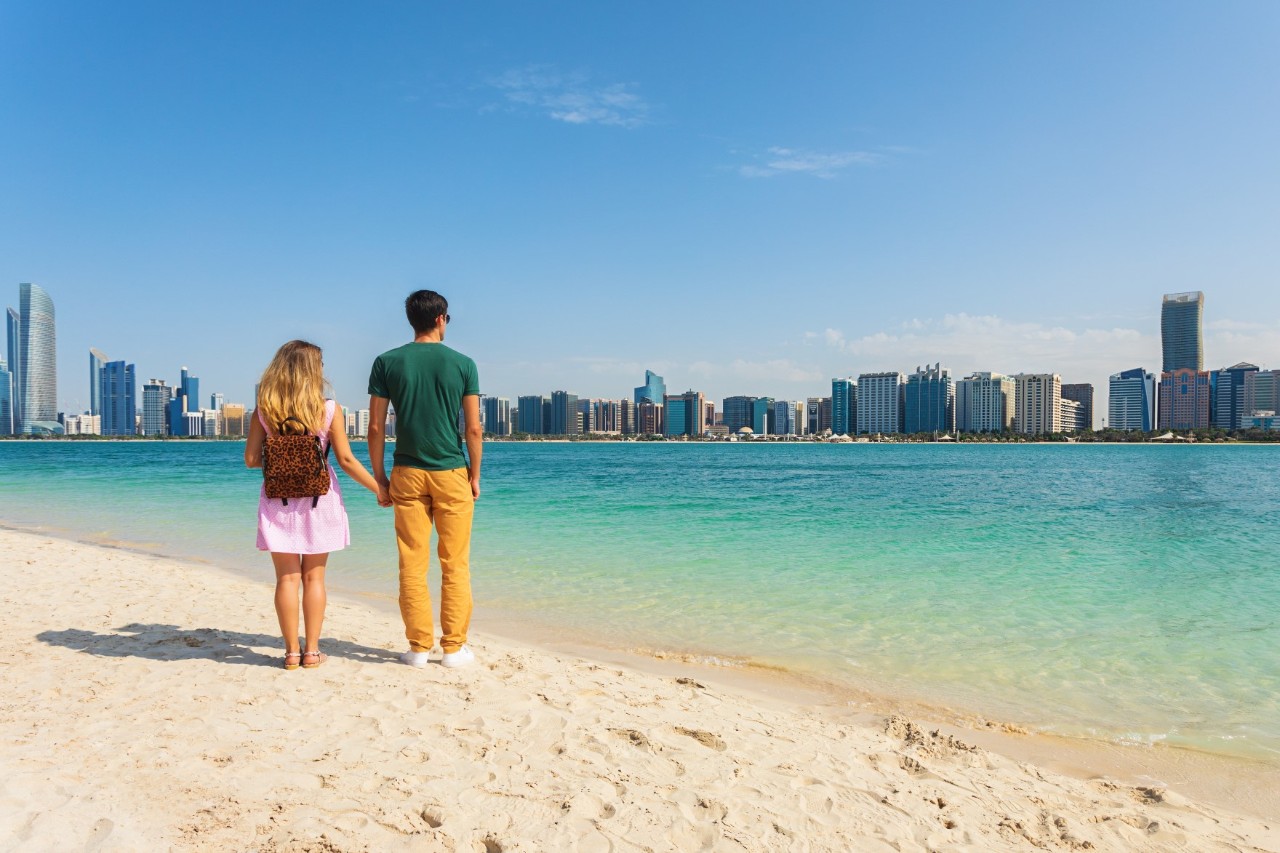 Ein Paar steht an einem Strand und blickt über das türkisgrüne Wasser einer Bucht auf eine große Stadt mit modernen Hochhäusern.