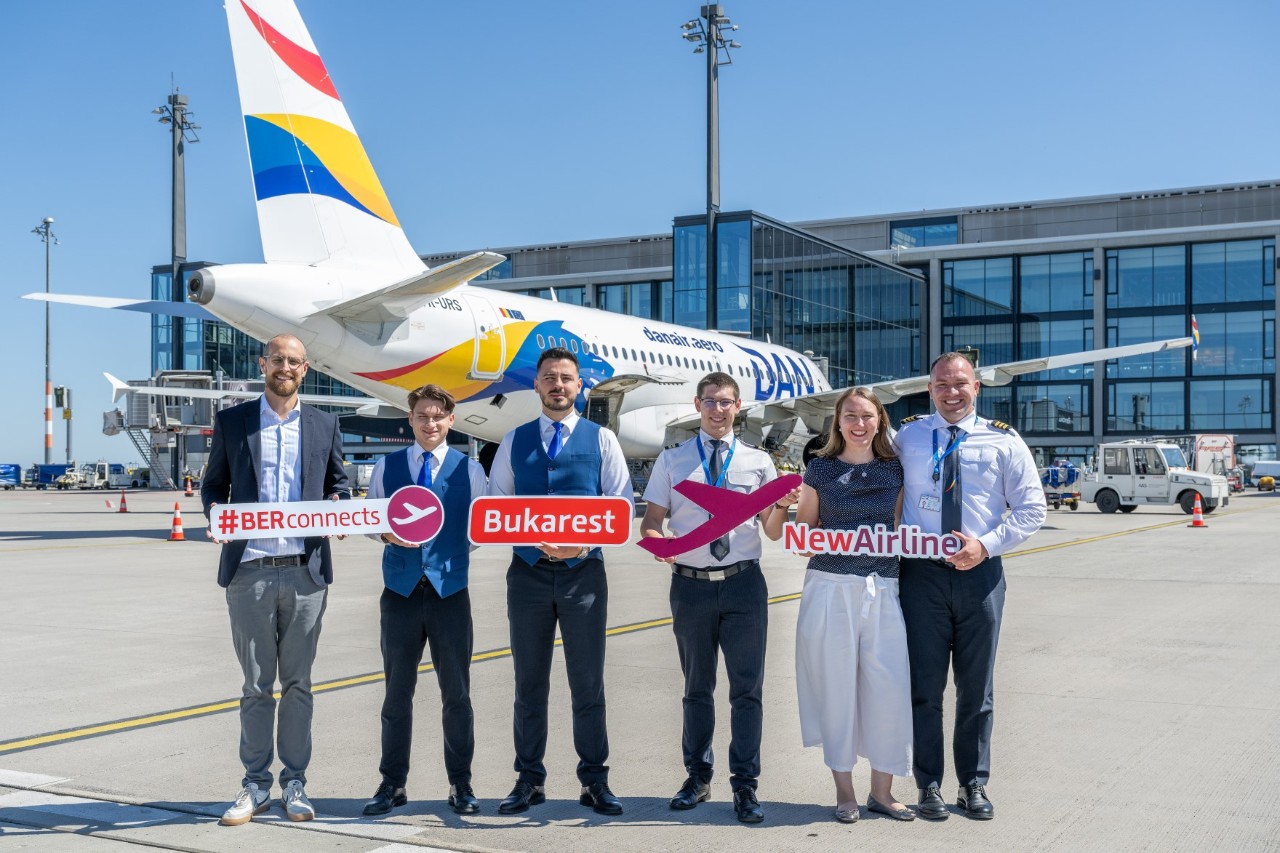 Sechs Personen stehen vor einem Flugzeug auf dem Vorfeld. Sie posieren lächelnd und halten Schilder mit verschiedenen Aufschriften, darunter das Wort "Bukarest", und Flugzeug-Piktogrammen in den Händen. Einige der Personen tragen die Uniformen einer Flugzeug-Crew.