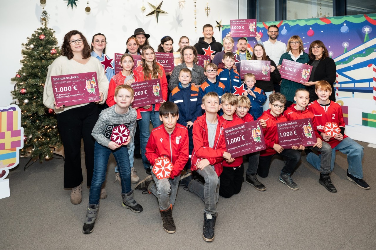 Eine Gruppe von Kindern und Erwachsenen posiert in einem weihnachtlich dekorierten Raum mit großen symbolischen Spendenchecks.